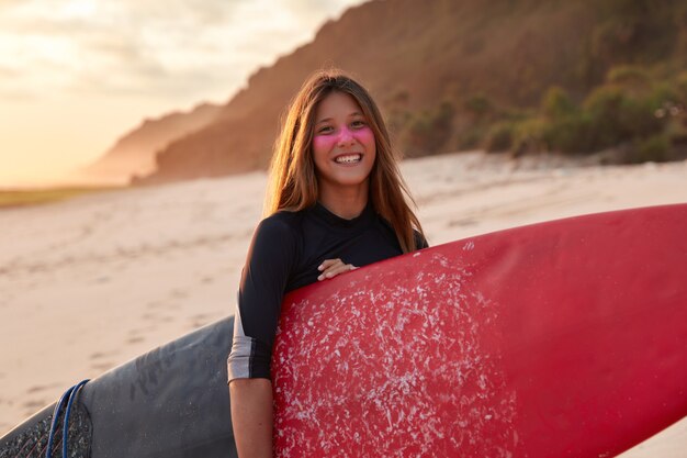 La deportista tiene entrenamiento en la playa, lleva una tabla de remo, sonríe con alegría, usa traje de neopreno, tiene una máscara protectora de zinc en la cara, posa cerca del arrecife