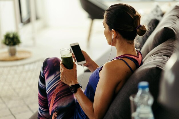 Deportista usando teléfono móvil y bebiendo batido de desintoxicación en el sofá en casa