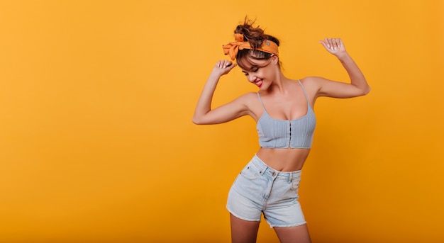 Deportiva chica encantadora en camiseta sin mangas y pantalones cortos de mezclilla bailando con placer y sonriendo