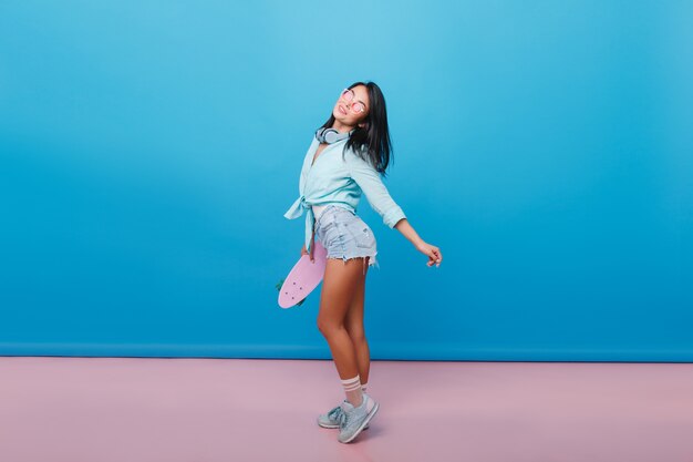 Deportiva chica hispana de pelo castaño en traje de mezclilla mirando hacia arriba. Maravillosa chica latina en elegantes zapatos con longboard