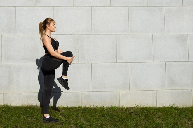 Deportiva chica rubia con cola de caballo y entrenamiento de cuerpo atlético en el parque por la mañana, estirando los músculos de las piernas después del entrenamiento cardiovascular, posando sobre fondo de pared de ladrillo gris con espacio de copia para su texto