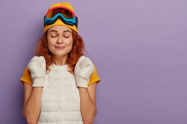 Deportiva joven disfruta de la estación de esquí, aprieta los puños con triunfo, mantiene los ojos cerrados, usa gafas protectoras de snowboard, aisladas sobre la pared púrpura del estudio.