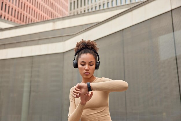 Deportiva joven de pelo rizado se encuentra al aire libre descansa después de que el entrenamiento comprueba los resultados en el reloj inteligente escucha música en los auriculares lleva ropa activa lleva un estilo de vida deportivo. Concepto de dispositivos de fitness