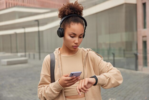 Deportiva mujer con cabello rizado revisa calorías en smartwatch usa teléfono móvil escucha música a través de auriculares vestida con ropa deportiva se encuentra al aire libre contra un fondo borroso Estilo de vida deportivo