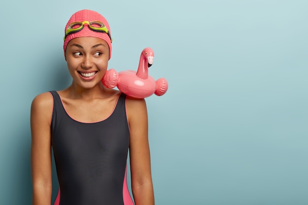 Deportiva mujer delgada con piel oscura viste traje de baño, tiene un anillo de natación en forma de flamenco rosa, gafas en la cabeza, pasa el tiempo libre en un centro de ocio contemporáneo, lista para nadar. Descanso activo