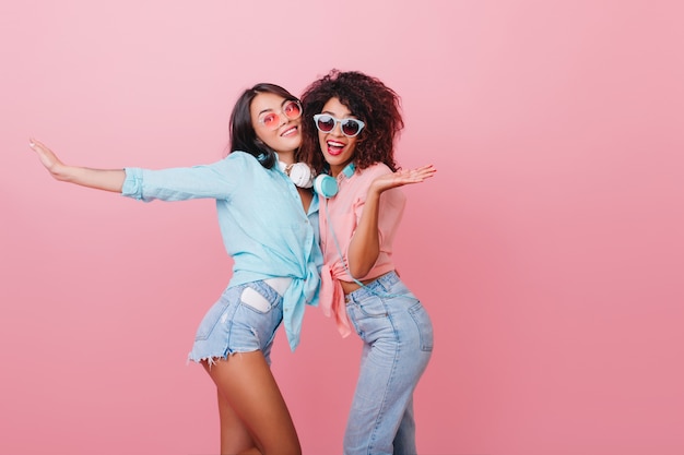 Foto gratuita deportiva mujer europea con piel bronceada bailando con alegre amigo africano. felices dos chicas en atuendos de verano jugando juntas.