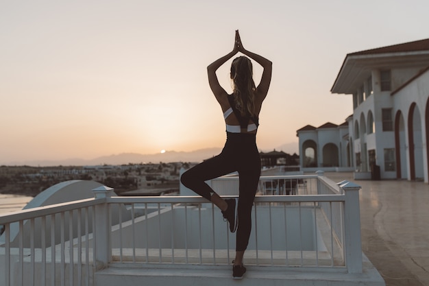 Deportiva mujer joven atractiva relajante formación de yoga al amanecer en el paseo marítimo. Ver la espalda, el trabajo al aire libre, el estilo de vida deportivo, el estado de ánimo alegre, relajarse cerca del mar, entrenar al aire libre, verdaderas emociones positivas.