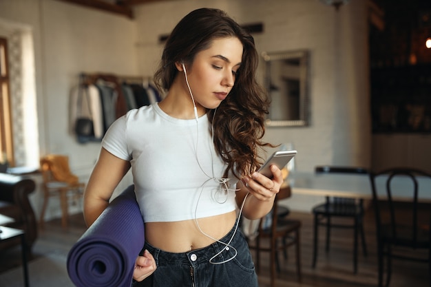 Deportiva mujer joven bronceada de talla grande en jeans y camiseta ajustada sosteniendo un tapete de fitness enrollado debajo del brazo, escribiendo un mensaje de texto en el teléfono celular