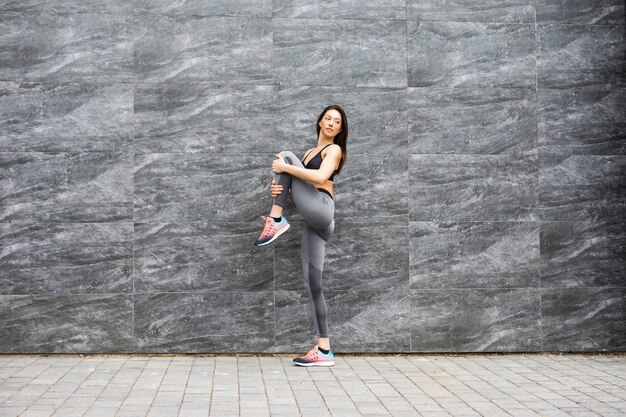 Deportiva mujer joven practicando yoga trabajando, vistiendo ropa deportiva, exterior de cuerpo entero, pared de ladrillo