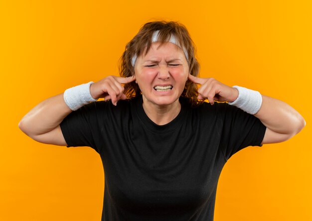 Deportiva mujer de mediana edad en camiseta negra con diadema cerrando las orejas con los dedos con expresión molesta de pie sobre la pared naranja