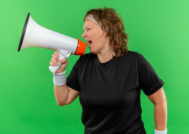 Deportiva mujer de mediana edad en camiseta negra con diadema gritando al megáfono con expresión agresiva de pie sobre la pared verde