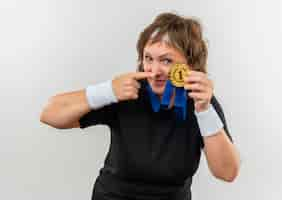 Foto gratuita deportiva mujer de mediana edad en camiseta negra con diadema y medalla de oro alrededor de su cuello apuntando con el dedo a ella sonriendo confiada de pie sobre la pared blanca