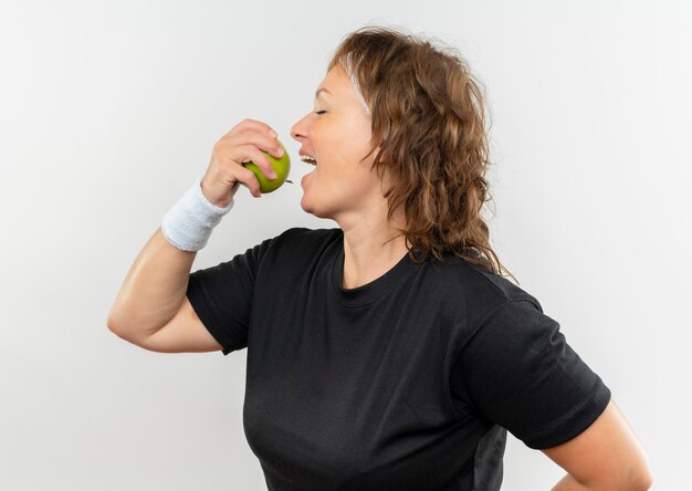 Deportiva mujer de mediana edad en camiseta negra con diadema sosteniendo manzana verde mordiéndola con cara feliz de pie sobre la pared blanca