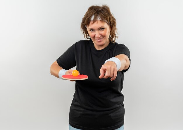 Deportiva mujer de mediana edad en camiseta negra con diadema sosteniendo raqueta con pelota de tenis de mesa apuntando con el dedo a la cámara sonriendo con cara feliz de pie sobre la pared blanca