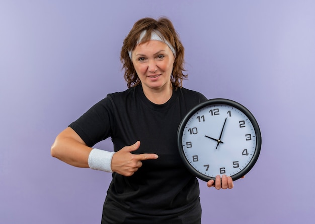 Deportiva mujer de mediana edad en camiseta negra con diadema sosteniendo reloj de pared apuntando con el dedo a él sonriendo confiado de pie sobre la pared azul