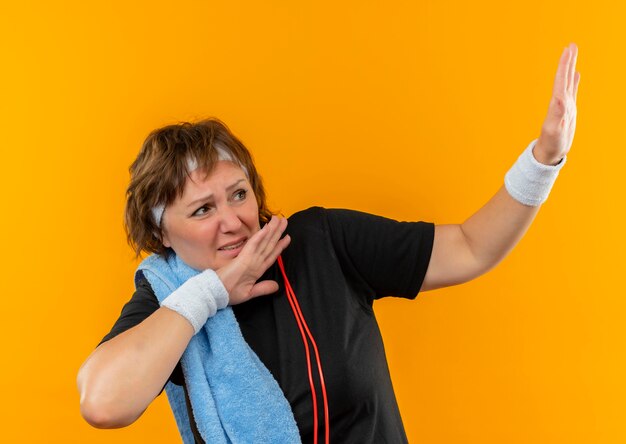 Deportiva mujer de mediana edad en camiseta negra con diadema y con una toalla en el hombro haciendo un gesto de defensa con expresión de miedo en la cara de pie sobre la pared naranja