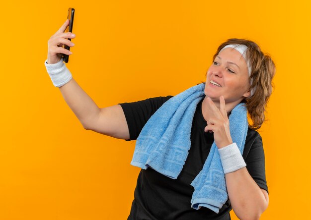 Deportiva mujer de mediana edad en camiseta negra con diadema y con toalla en el hombro oh su teléfono inteligente tomando selfie sonriendo con cara feliz de pie sobre la pared naranja