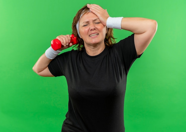 Deportiva mujer de mediana edad en camiseta negra con diadema trabajando con mancuernas mirando cansado y agotado de pie sobre la pared verde