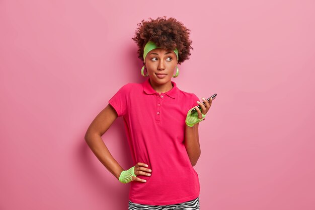 Deportiva mujer morena de pelo rizado mantiene la mano en la cintura, sostiene el teléfono móvil en la mano, usa una aplicación especial para entrenar en línea, usa diadema, guantes deportivos, camiseta rosa