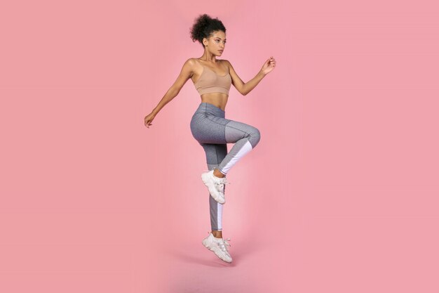 Deportiva mujer practicando ejercicios de sentadillas en estudio. Mujer africana en ropa deportiva trabajando sobre fondo rosa.