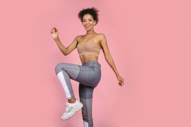 Deportiva mujer practicando ejercicios de sentadillas en estudio. Mujer africana en ropa deportiva trabajando sobre fondo rosa.