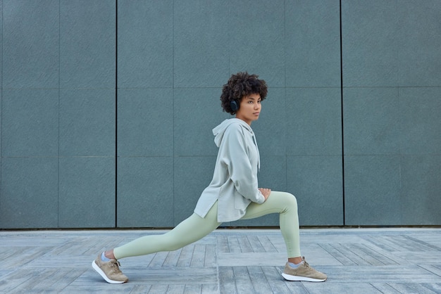 Deportiva mujer rizada estira las piernas y se calienta antes de que el entrenamiento se concentre pensativamente, usa leggings de sudadera y zapatillas de deporte posa contra la pared gris