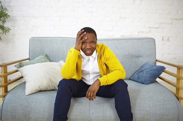 Deprimido joven afroamericano vestido informalmente sentado en el sofá en casa, sosteniendo la mano en la cabeza, viendo el campeonato de fútbol, sintiéndose molesto mientras su equipo favorito está perdiendo el juego