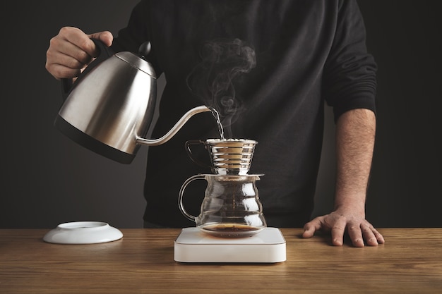 Derrama agua caliente para preparar café filtrado desde una tetera plateada hasta una hermosa cafetera de goteo cromado transparente sobre pesas blancas simples.