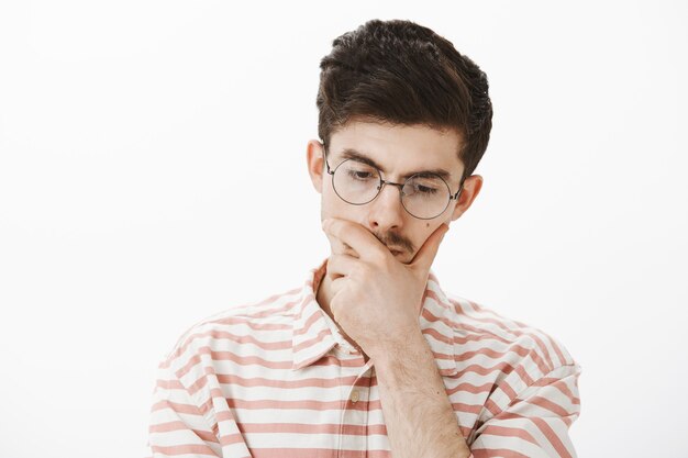 Desarrollador de programa masculino de pensamiento enfocado con gafas de moda, inclinando la cabeza hacia abajo y tocando la barbilla, preocupándose al tomar una decisión o planificar algo sobre una pared gris