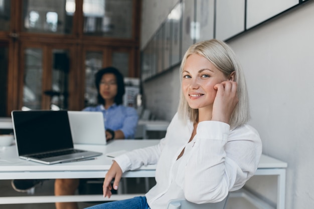 Desarrolladora web femenina posando con una sonrisa en la oficina mientras su colega asiático trabaja en el proyecto. Comercializador chino usando laptop sentado en la mesa con un gerente bastante europeo.