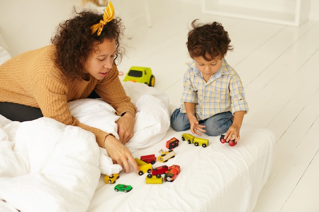 Desarrollo temprano, niñez y maternidad. Filmación en interiores de un niño pequeño curioso lindo sentado en el suelo jugando con muchos coches de juguete de colores en la sábana blanca, la madre junto a él, mirándolo jugar