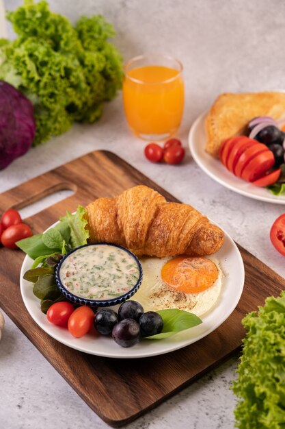 El desayuno consiste en croissant, huevo frito, aderezo para ensalada, uvas negras y tomates.