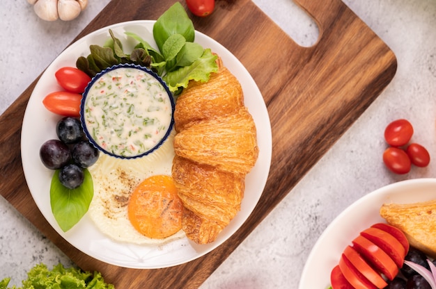 El desayuno consiste en croissant, huevo frito, aderezo para ensalada, uvas negras y tomates.