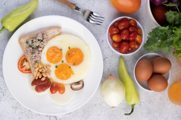 El desayuno consiste en huevos fritos, salchichas, carne de cerdo picada, pan, frijoles rojos y soja en un plato blanco.