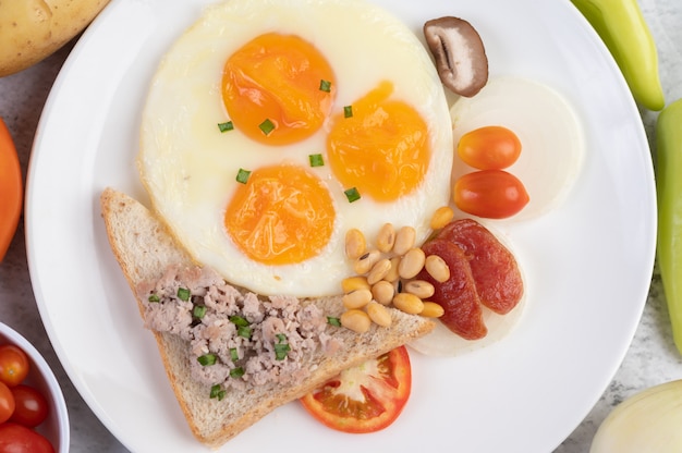 El desayuno consiste en huevos fritos, salchichas, carne de cerdo picada, pan, frijoles rojos y soja en un plato blanco.