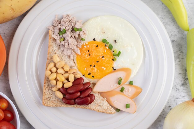 El desayuno consiste en huevos fritos, salchichas, carne de cerdo picada, pan, frijoles rojos y soja en un plato blanco.