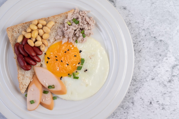 El desayuno consiste en huevos fritos, salchichas, carne de cerdo picada, pan, frijoles rojos y soja en un plato blanco.