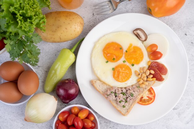 El desayuno consiste en huevos fritos, salchichas, carne de cerdo picada, pan, frijoles rojos y soja en un plato blanco.