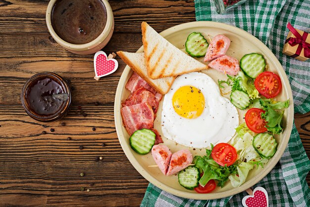 Desayuno en el Día de San Valentín: huevo frito en forma de corazón, tostadas, salchichas, tocino y verduras frescas. Desayuno inglés. Taza de cafe. Vista superior