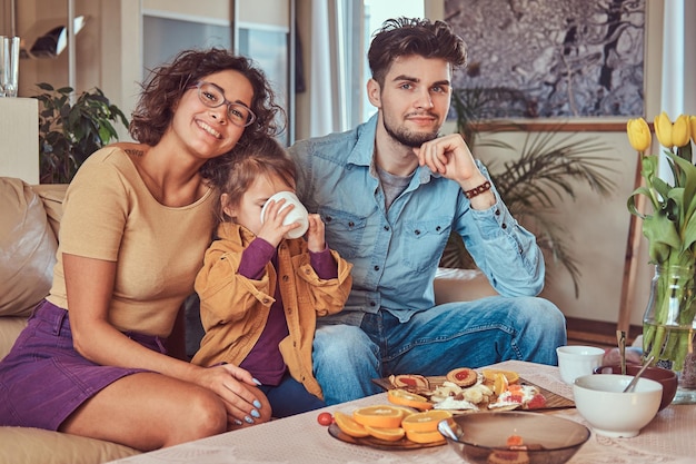 Desayuno familiar feliz. Joven familia atractiva desayunando en casa sentada en el sofá.