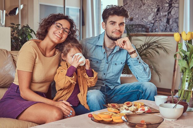 Desayuno familiar feliz. Joven familia atractiva desayunando en casa sentada en el sofá.