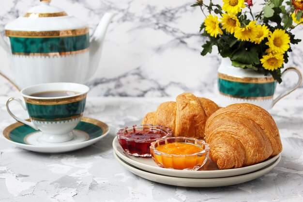 Desayuno francés con cruasanes, mermelada de albaricoque, mermelada de cereza y una taza de té, flores rojas y amarillas.