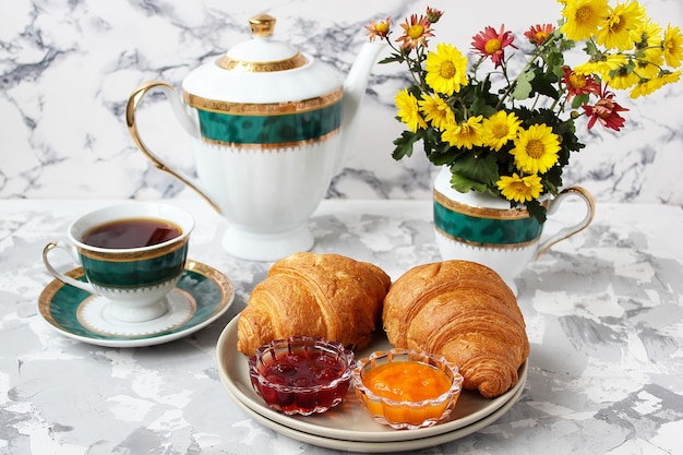 Desayuno francés con cruasanes, mermelada de albaricoque, mermelada de cereza y una taza de té, flores rojas y amarillas.