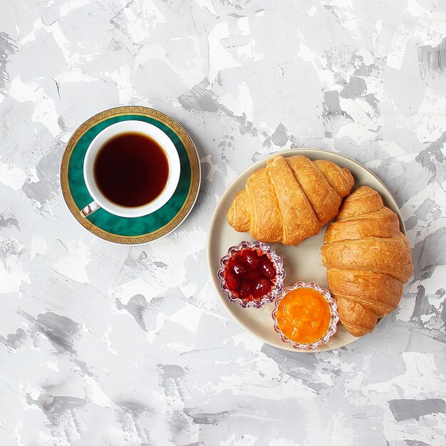 Desayuno francés con cruasanes, mermelada de albaricoque, mermelada de cereza y una taza de té, flores rojas y amarillas.