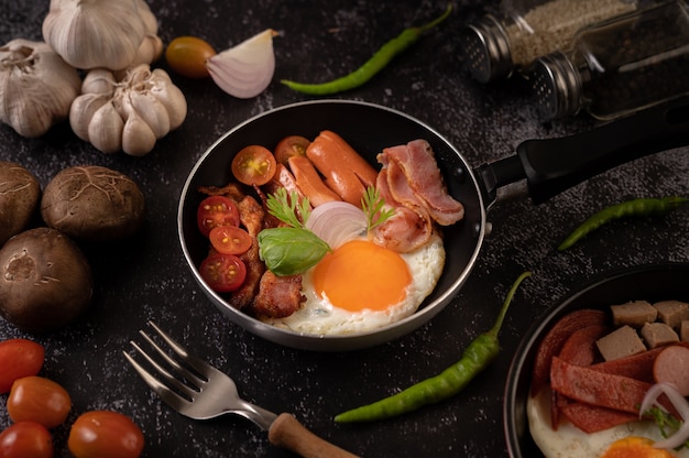 Desayuno con huevos fritos, salchicha y jamón en una sartén con tomates. Chile y albahaca