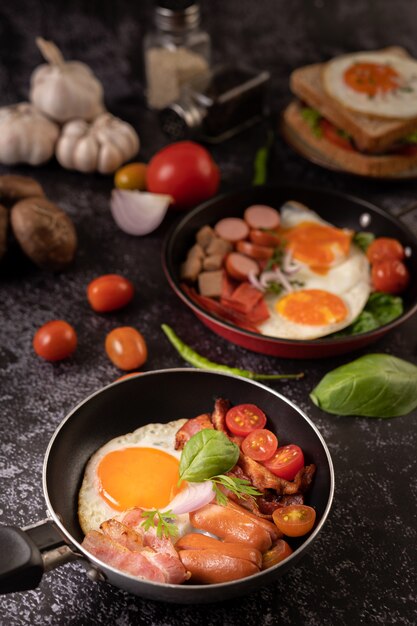 Desayuno con huevos fritos, salchicha y jamón en una sartén con tomates. Chile y albahaca