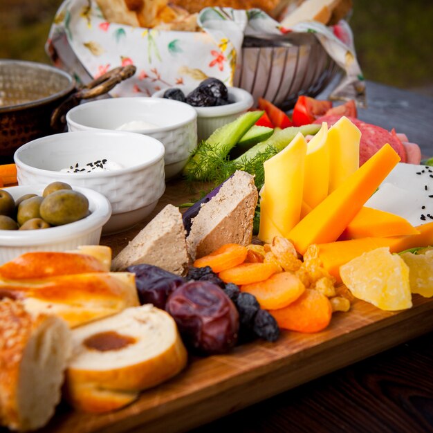 Desayuno en primer plano diferentes tipos de queso, pepinos, tomates, lechuga, albaricoques secos, pasas, dátiles en un soporte de madera