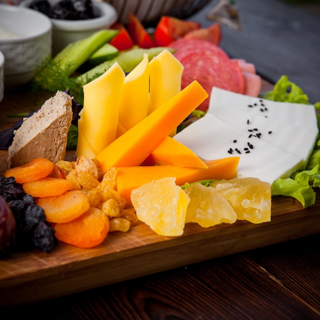 Desayuno en primer plano diferentes tipos de queso, pepinos, tomates, lechuga, albaricoques secos, pasas, dátiles en un soporte de madera