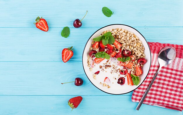 Desayuno saludable - granola, fresas, cerezas, nueces y yogurt en un recipiente sobre una mesa de madera. Concepto de comida vegetariana. Vista superior