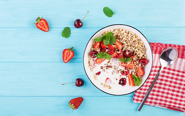 Desayuno saludable - granola, fresas, cerezas, nueces y yogurt en un recipiente sobre una mesa de madera. Concepto de comida vegetariana. Vista superior